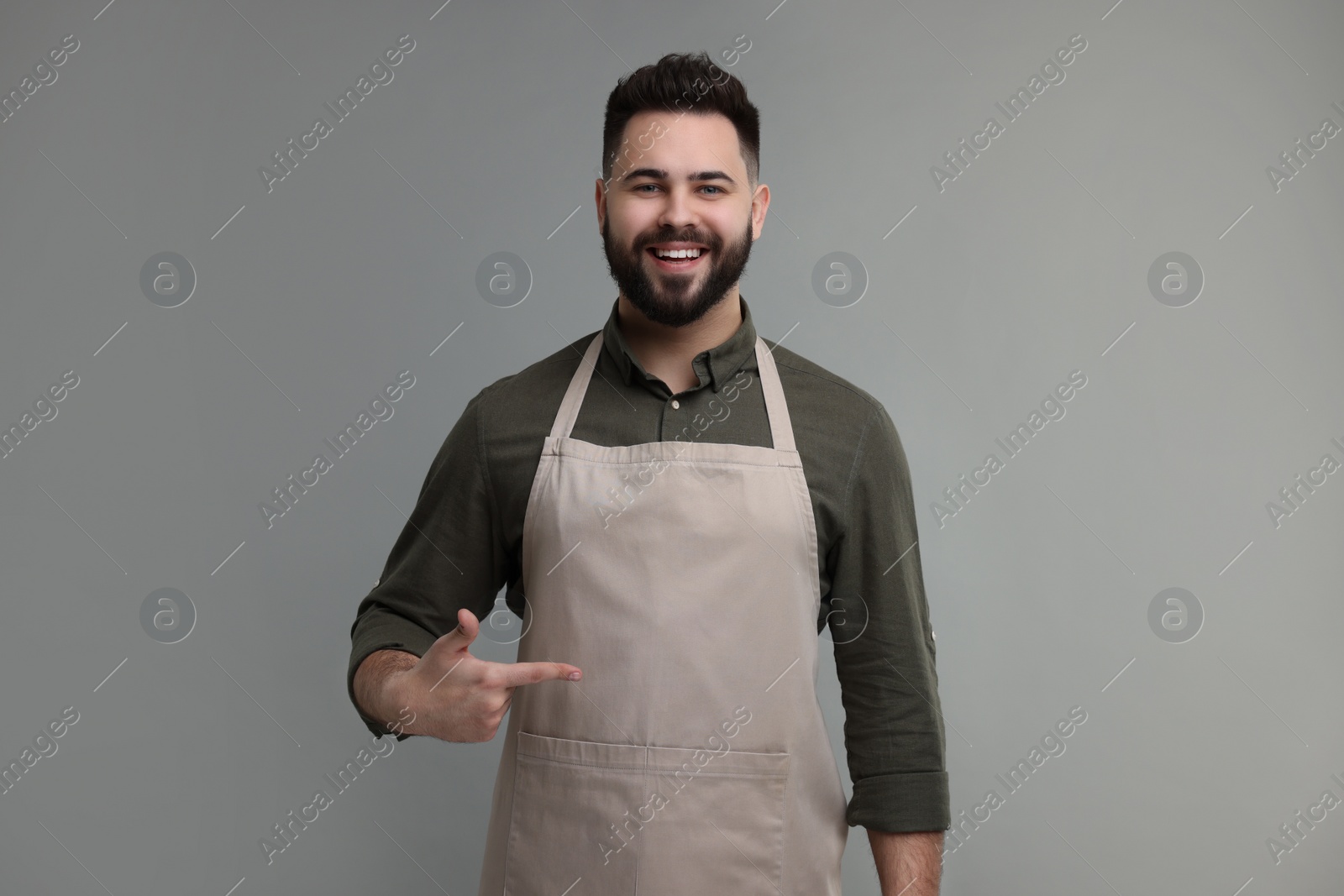 Photo of Smiling man pointing at kitchen apron on grey background. Mockup for design