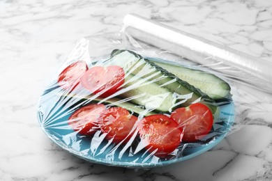 Photo of Plate of fresh vegetables with plastic food wrap at white marble table, closeup