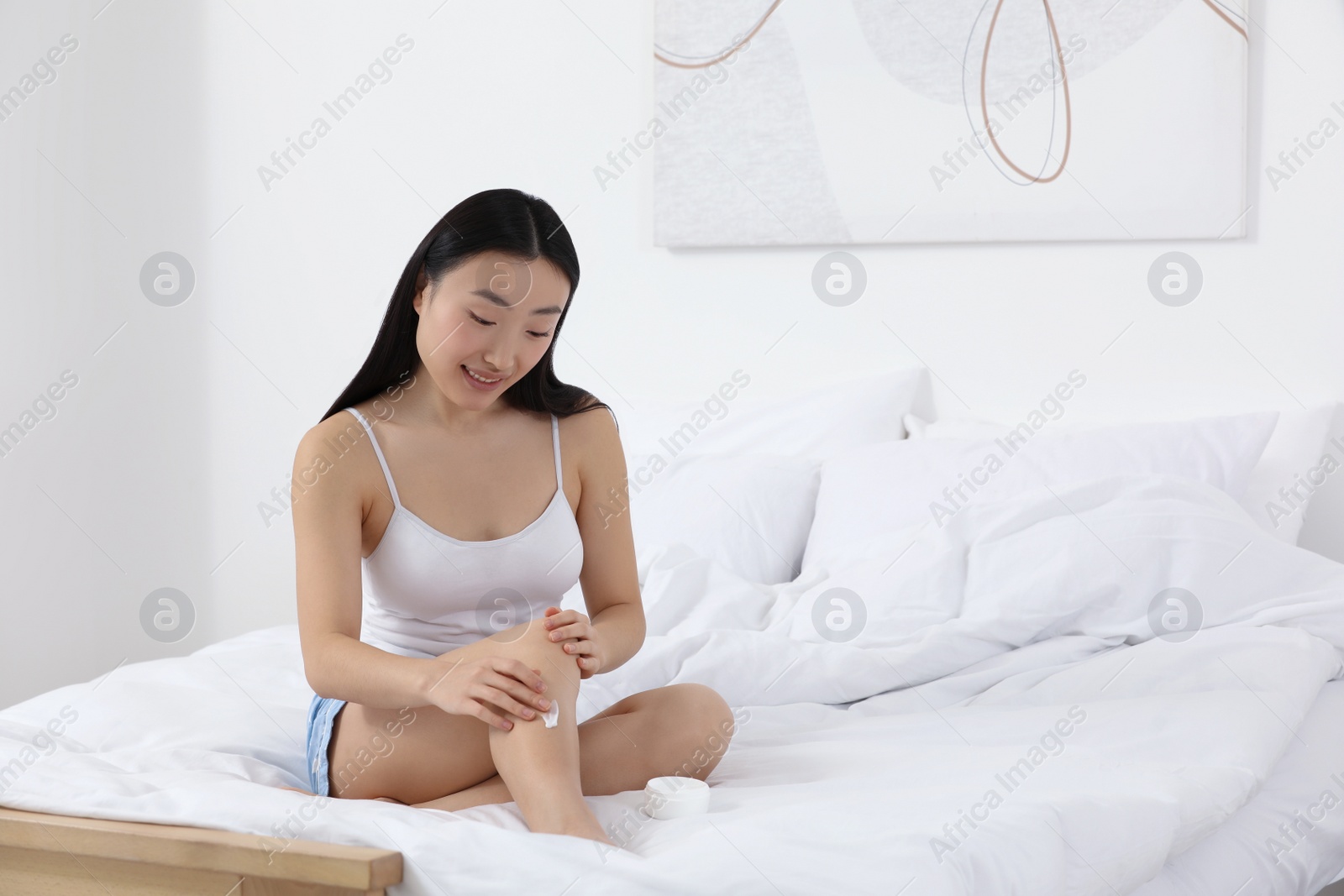 Photo of Beautiful young Asian woman applying body cream on leg in bedroom