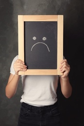 Woman hiding behind small chalkboard with drawing of sad face on dark background. Depression symptoms