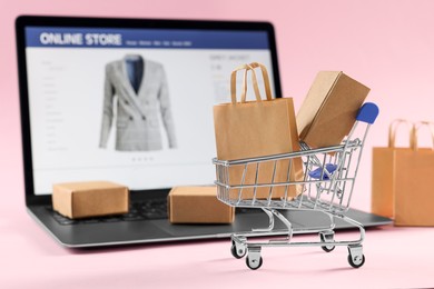Photo of Online store. Mini shopping cart, purchases and laptop on pink background, selective focus
