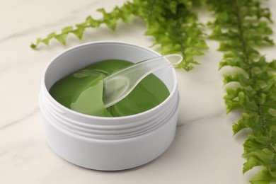 Photo of Jar of under eye patches with spoon and green fern on white marble table, closeup. Cosmetic product