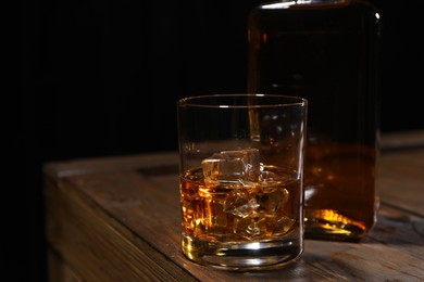Photo of Whiskey with ice cubes in glass and bottle on wooden crate against black background, closeup. Space for text