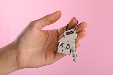 Woman holding key with metallic keychain in shape of house on pink background, closeup