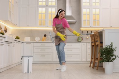 Photo of Enjoying cleaning. Happy woman in headphones dancing with mop in kitchen