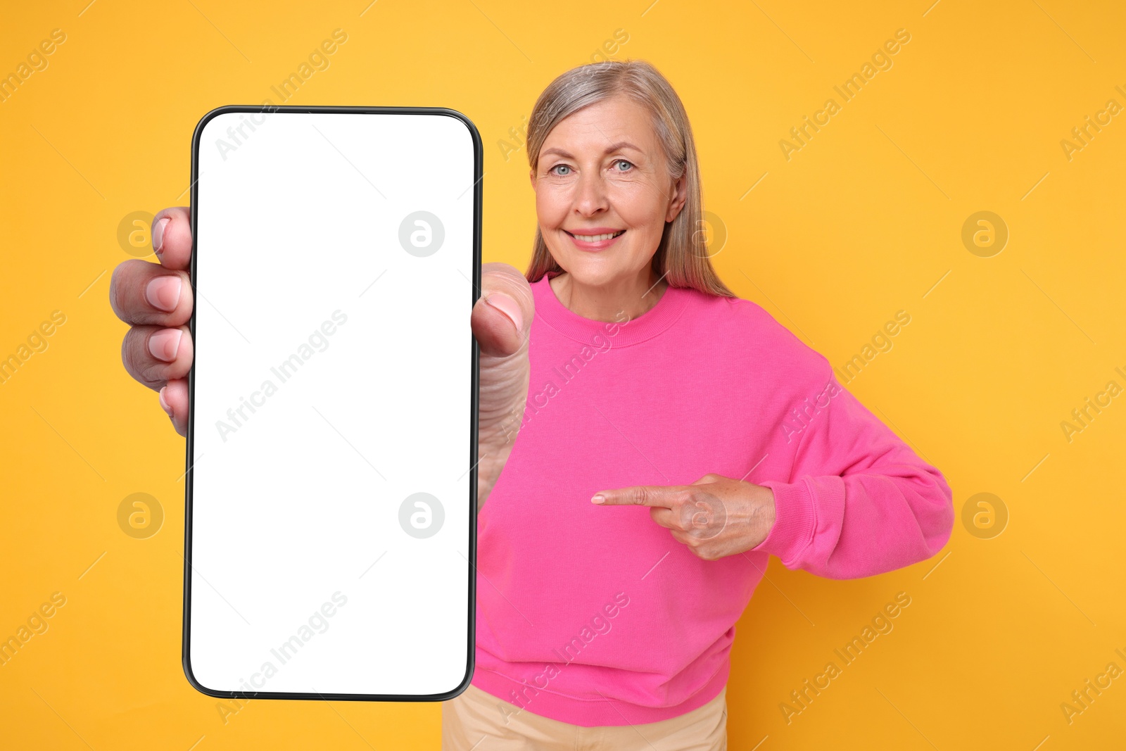 Image of Happy mature woman pointing at mobile phone with blank screen on golden background. Mockup for design