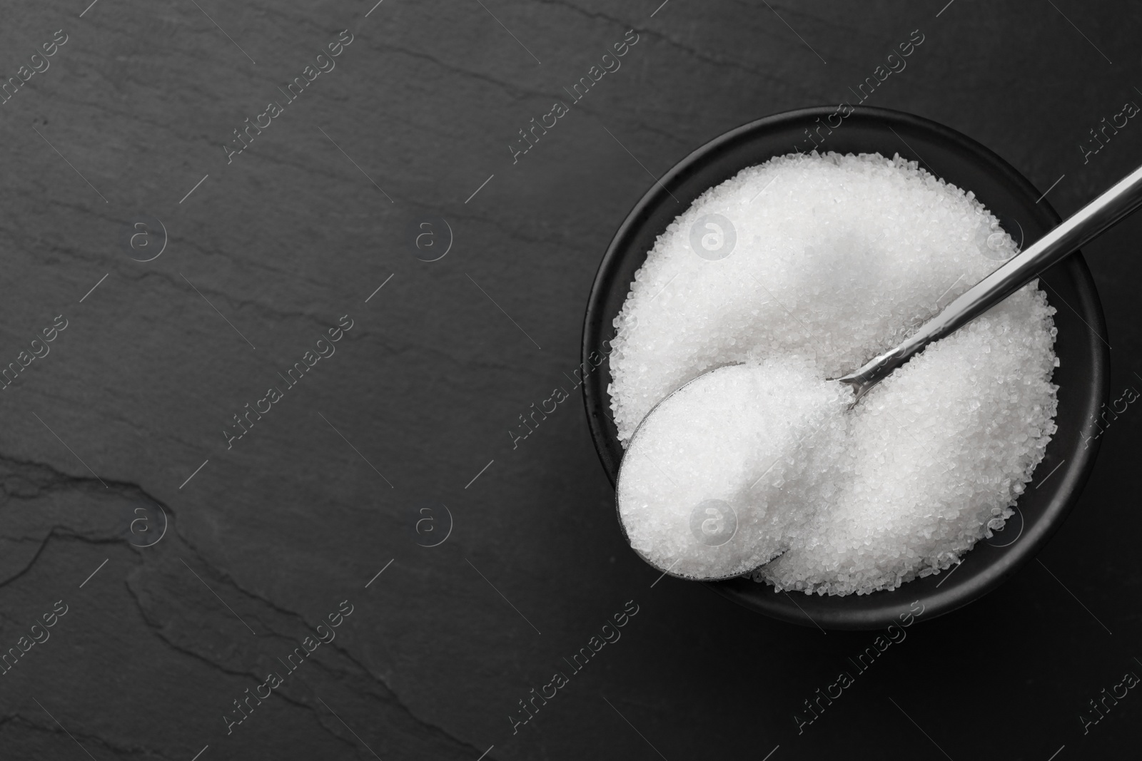 Photo of Granulated sugar and spoon in bowl on black table, top view. Space for text