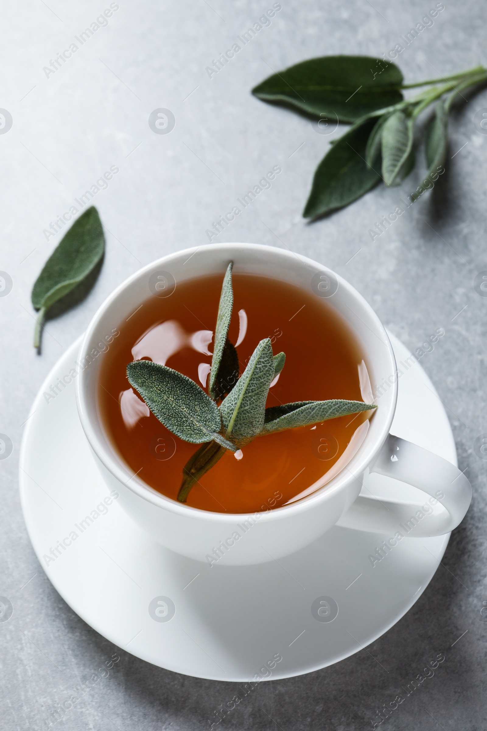 Photo of Cup of aromatic sage tea and fresh leaves on grey table