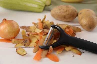 Photo of Peels of fresh vegetables and peeler on white wooden table, closeup