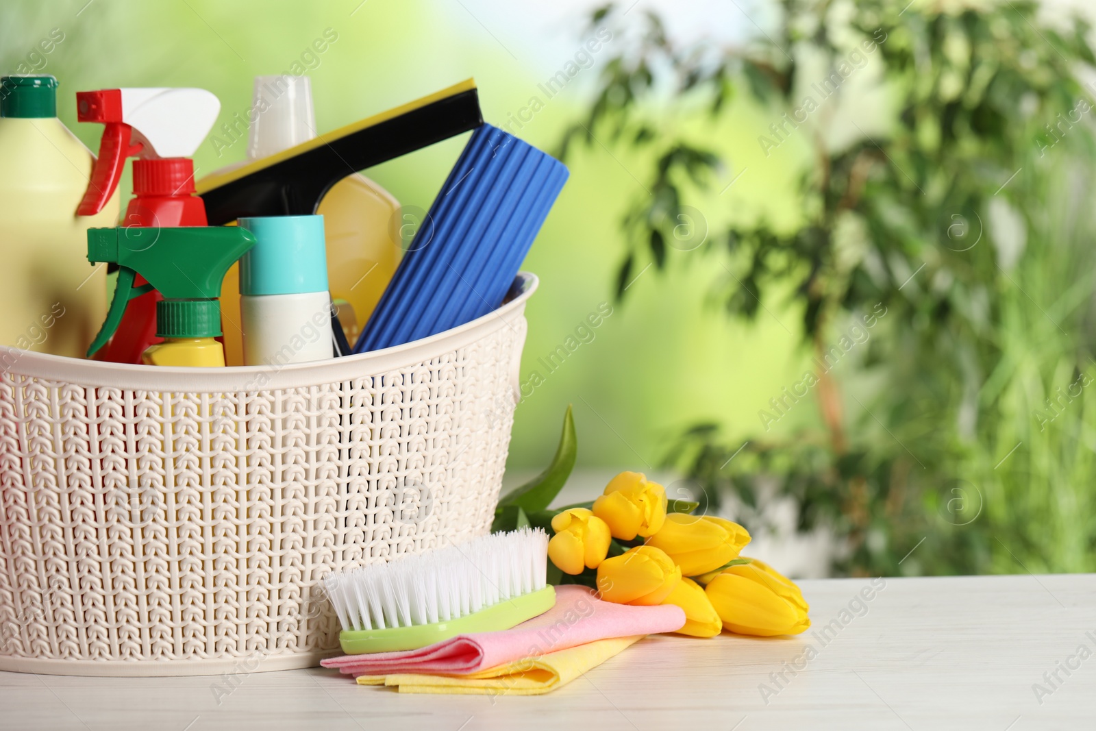 Photo of Spring cleaning. Plastic basket with detergents, supplies and beautiful flowers on white wooden table outdoors, space for text