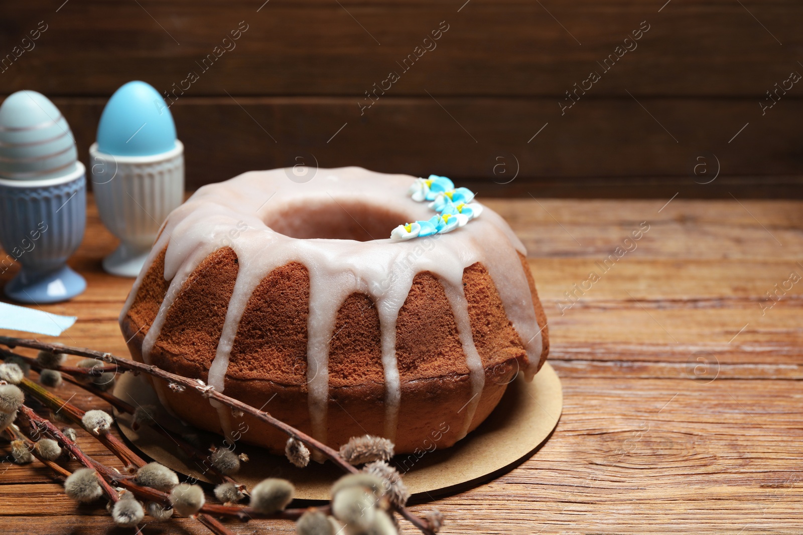 Photo of Delicious Easter cake decorated with sprinkles near willow branches and painted eggs on wooden table, closeup. Space for text