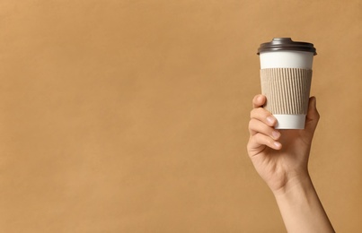 Woman holding takeaway paper coffee cup with cardboard sleeve on brown background, closeup. Space for text