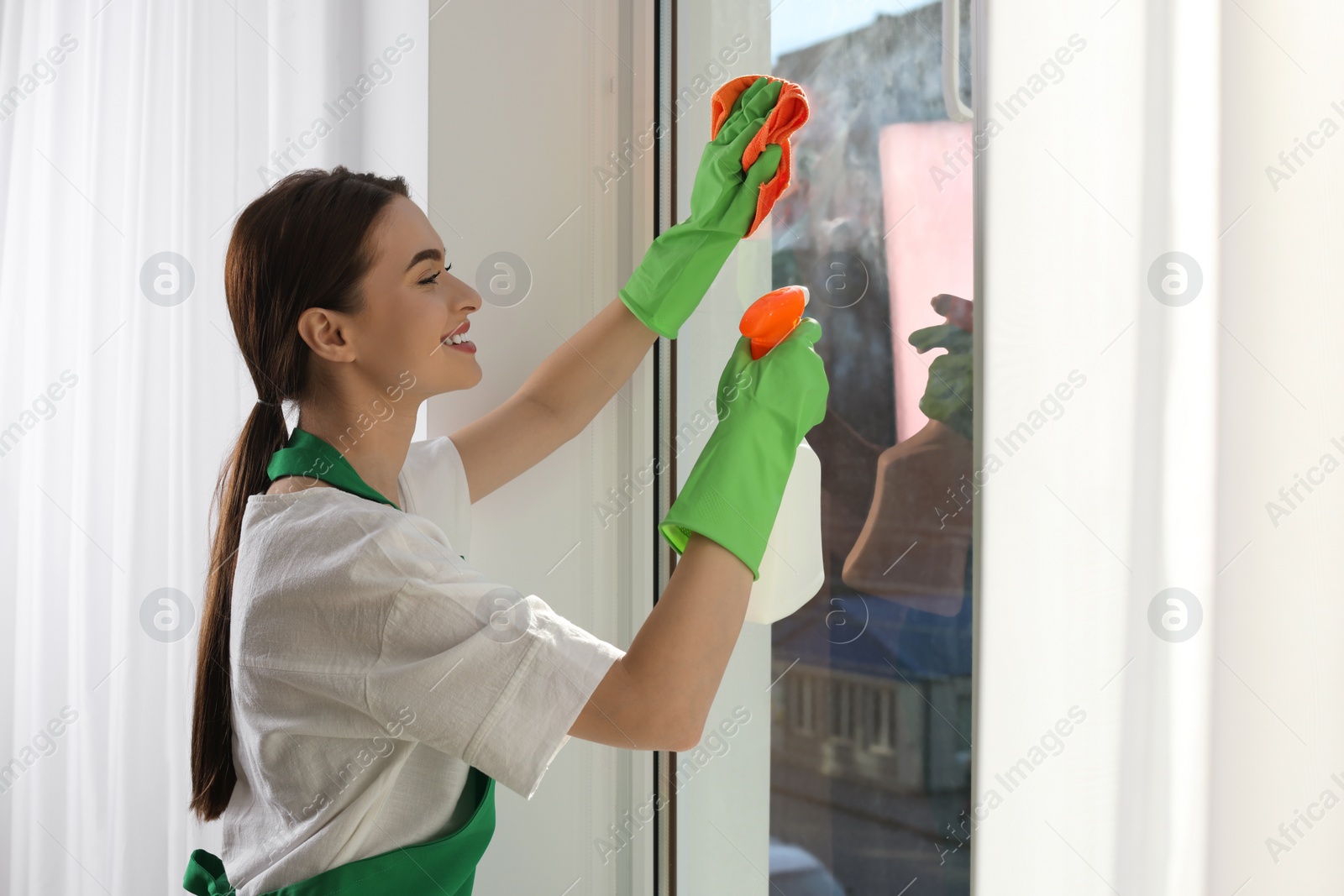 Photo of Young woman cleaning window glass with rag and detergent at home