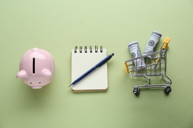 Notebook, piggy bank and cart with money on green background, flat lay