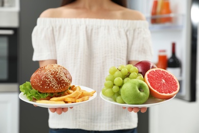 Concept of choice. Woman holding fruits and burger with French fries near refrigerator in kitchen, closeup