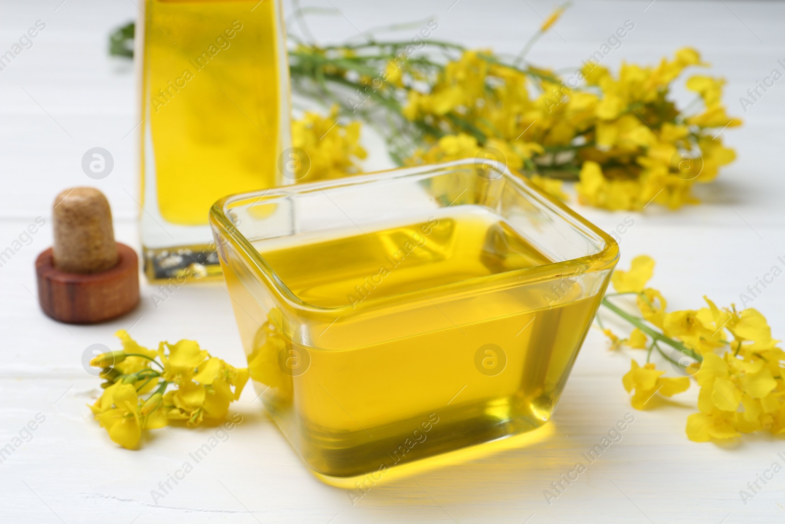 Photo of Rapeseed oil in glass bowl, bottle and beautiful yellow flowers on white wooden table, closeup