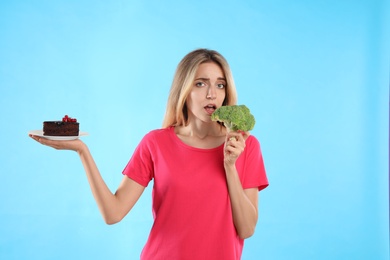 Woman choosing between cake and healthy broccoli on light blue background