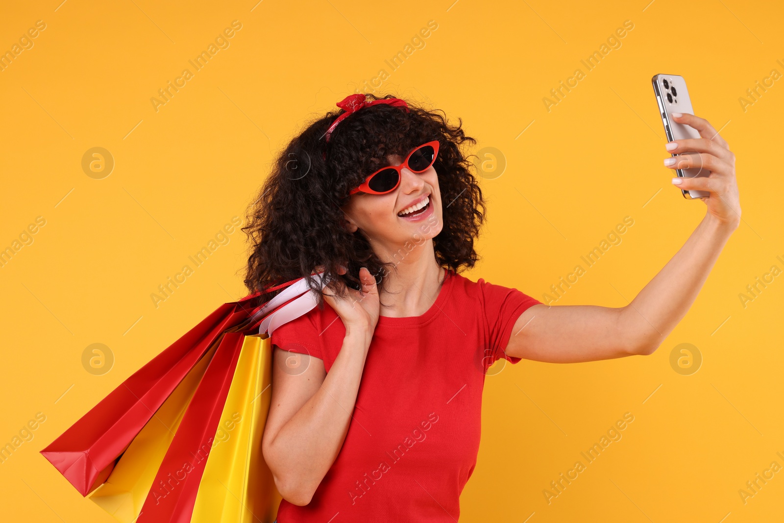 Photo of Happy young woman with shopping bags taking selfie on yellow background