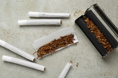 Photo of Roller with tobacco and hand rolled cigarettes on light grey table, flat lay