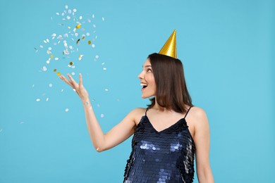 Happy young woman in party hat near flying confetti on light blue background