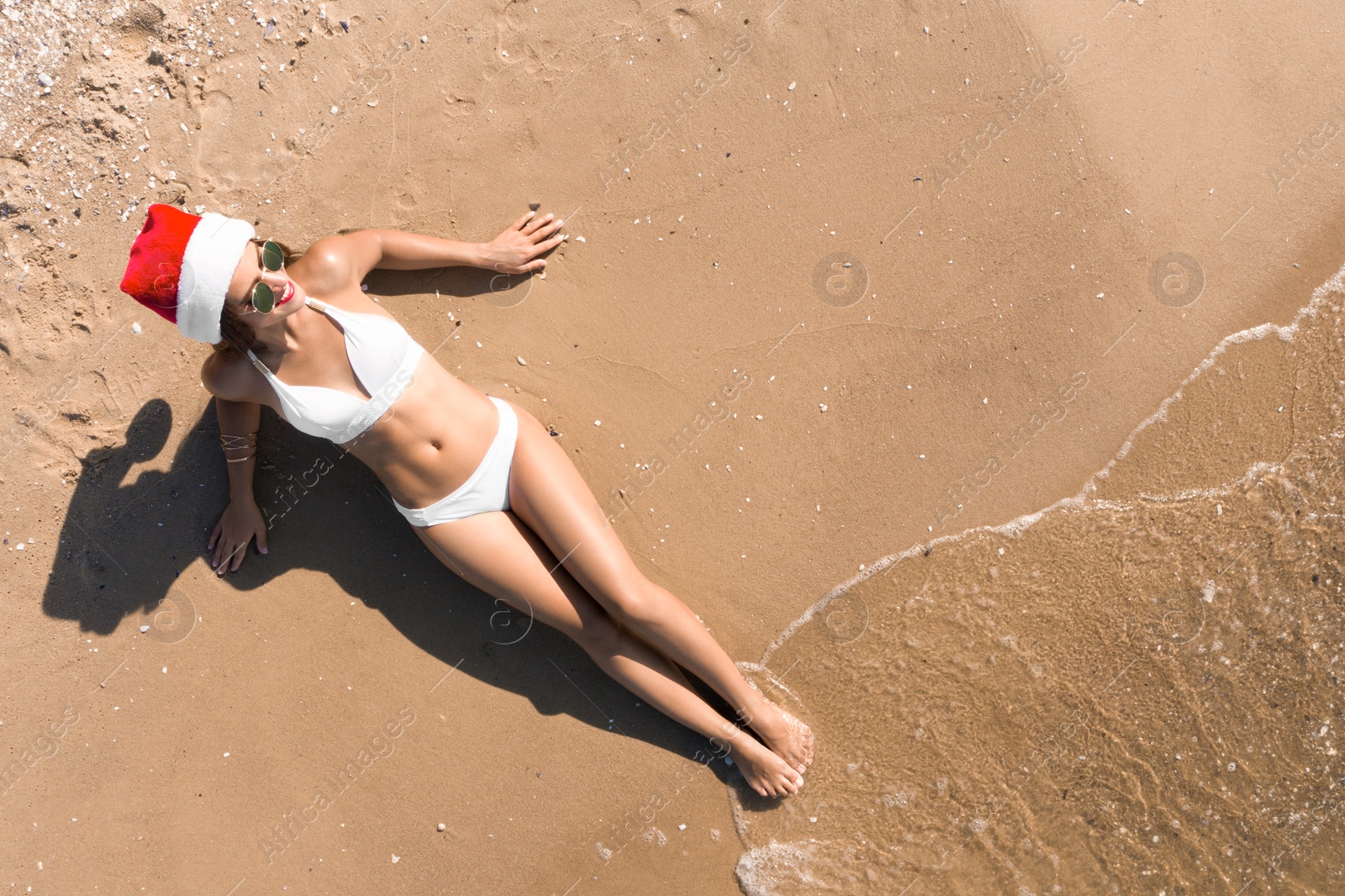 Image of Young woman wearing Santa hat and bikini on sunny beach, top view. Christmas vacation