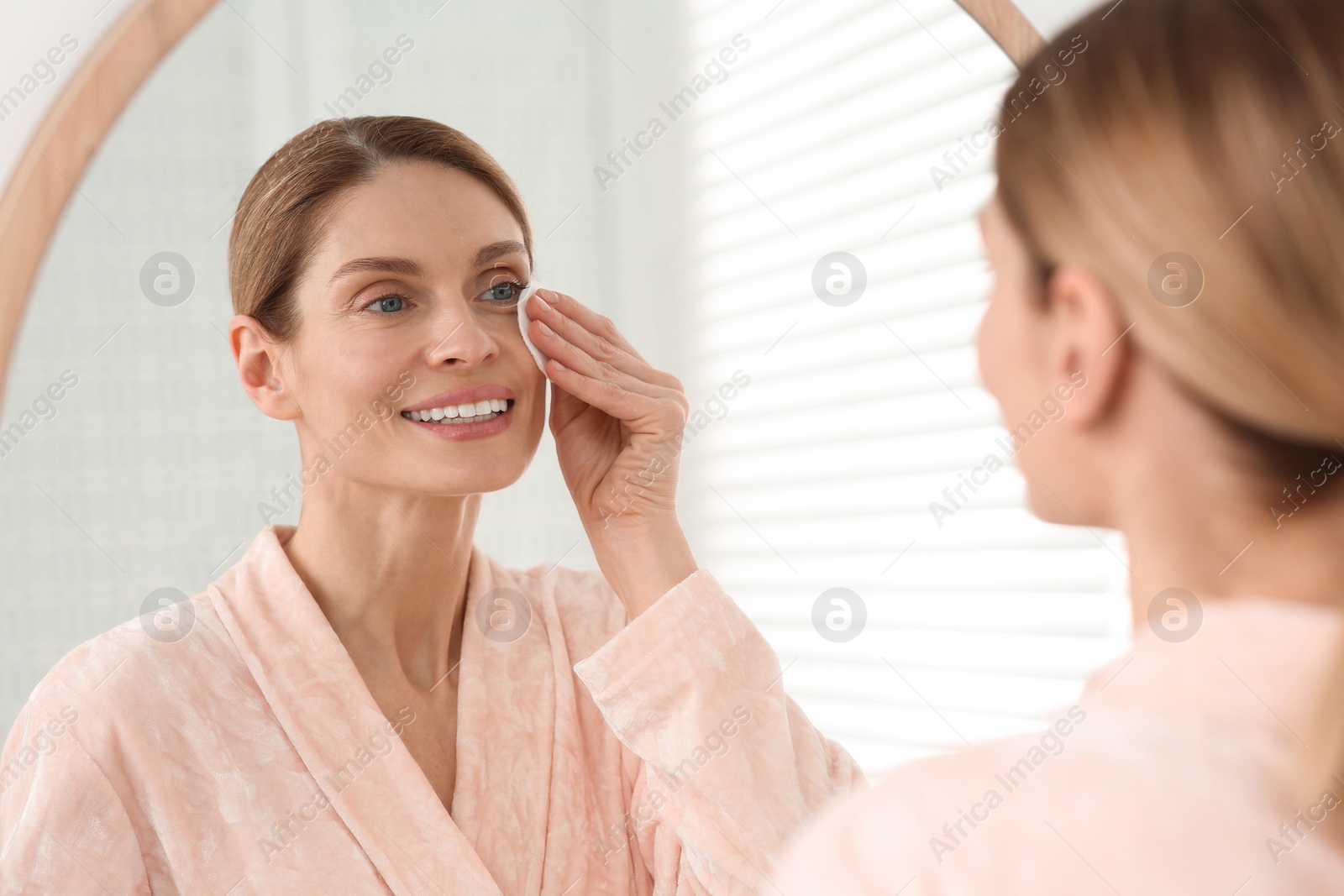 Photo of Beautiful woman removing makeup with cotton pad near mirror indoors