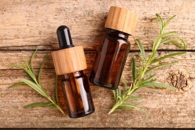 Aromatic essential oils in bottles and rosemary on wooden table, flat lay