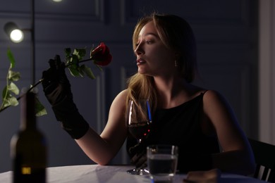 Photo of Elegant young woman with glass of wine and rose at table indoors in evening