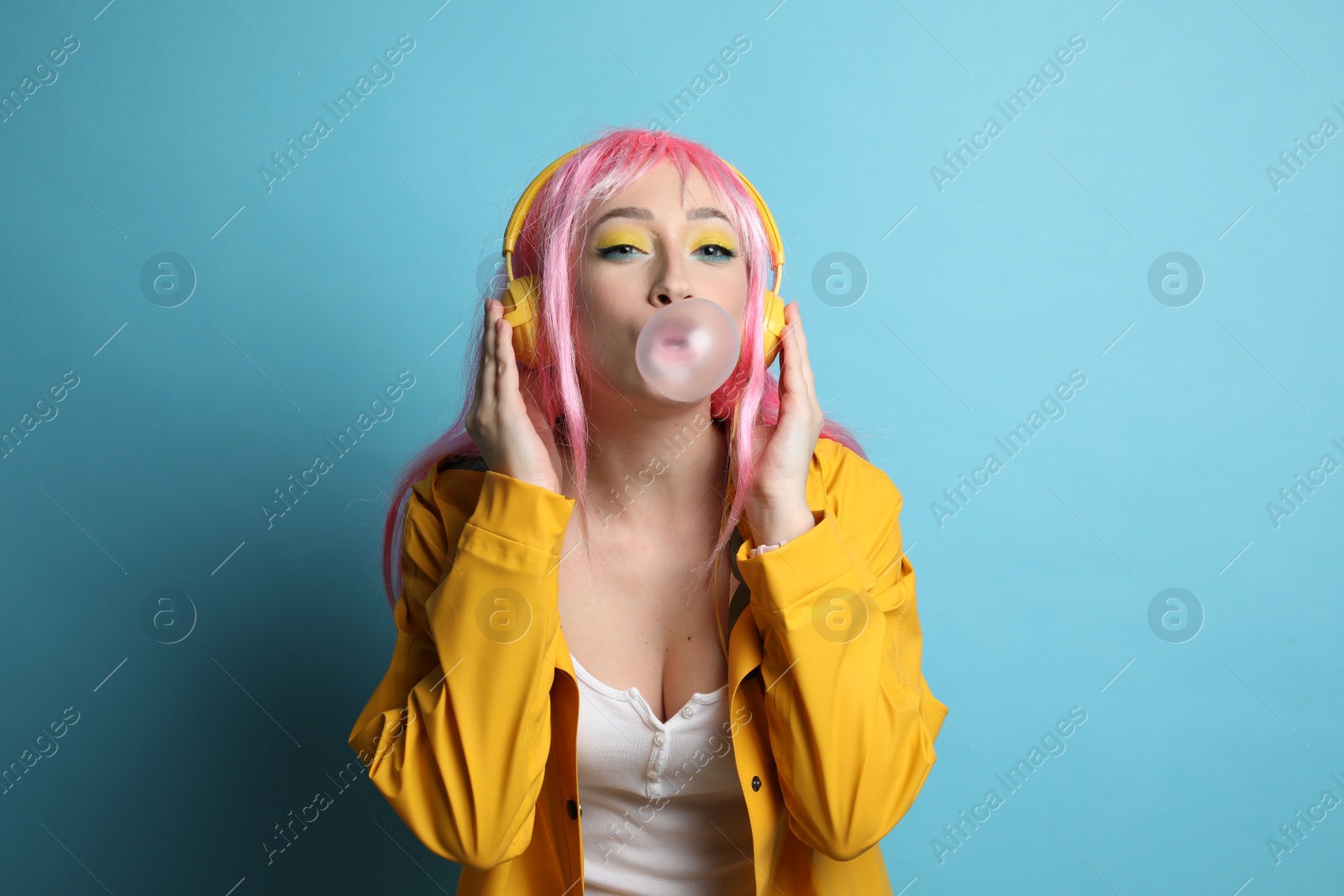 Photo of Fashionable young woman in pink wig with headphones blowing bubblegum on yellow background