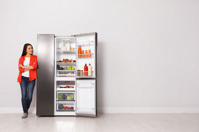 Photo of Happy young woman near open refrigerator indoors, space for text