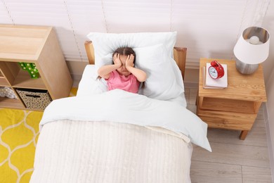 Photo of Little girl covering face in bed and alarm clock on bedside table at home, above view