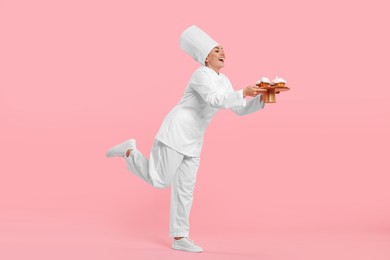 Photo of Happy professional confectioner in uniform holding cupcakes on pink background