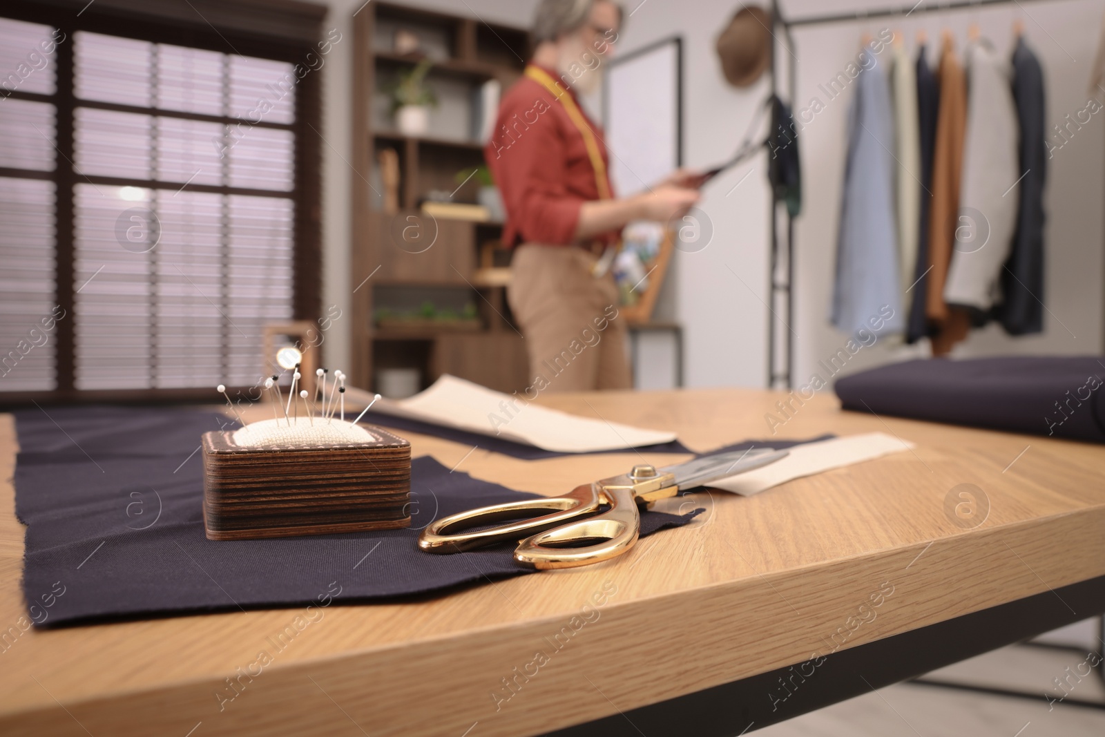 Photo of Professional tailor working in atelier, focus on table with scissors and pin cushion. Space for text