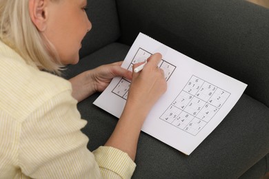 Photo of Middle aged woman solving sudoku puzzle on sofa at home, closeup