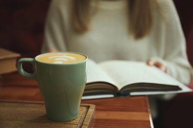 Woman with coffee reading book indoors, focus on cup