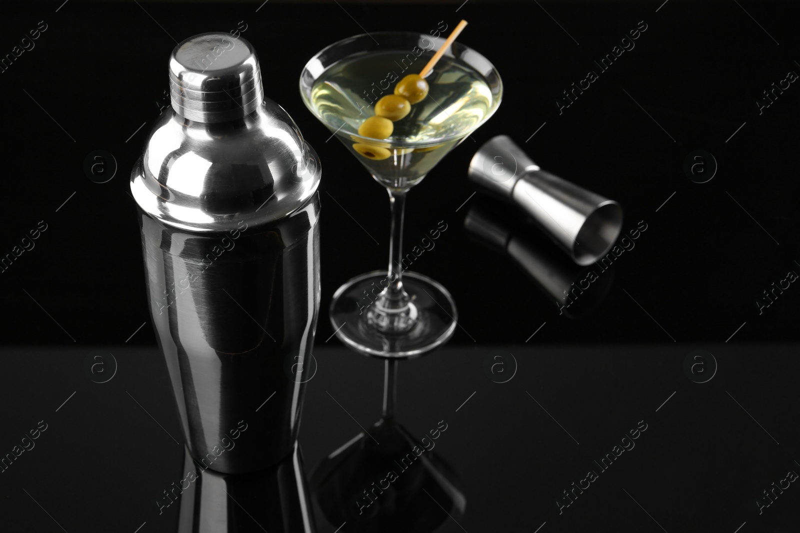 Photo of Metal shaker, Martini cocktail and jigger on black mirror surface