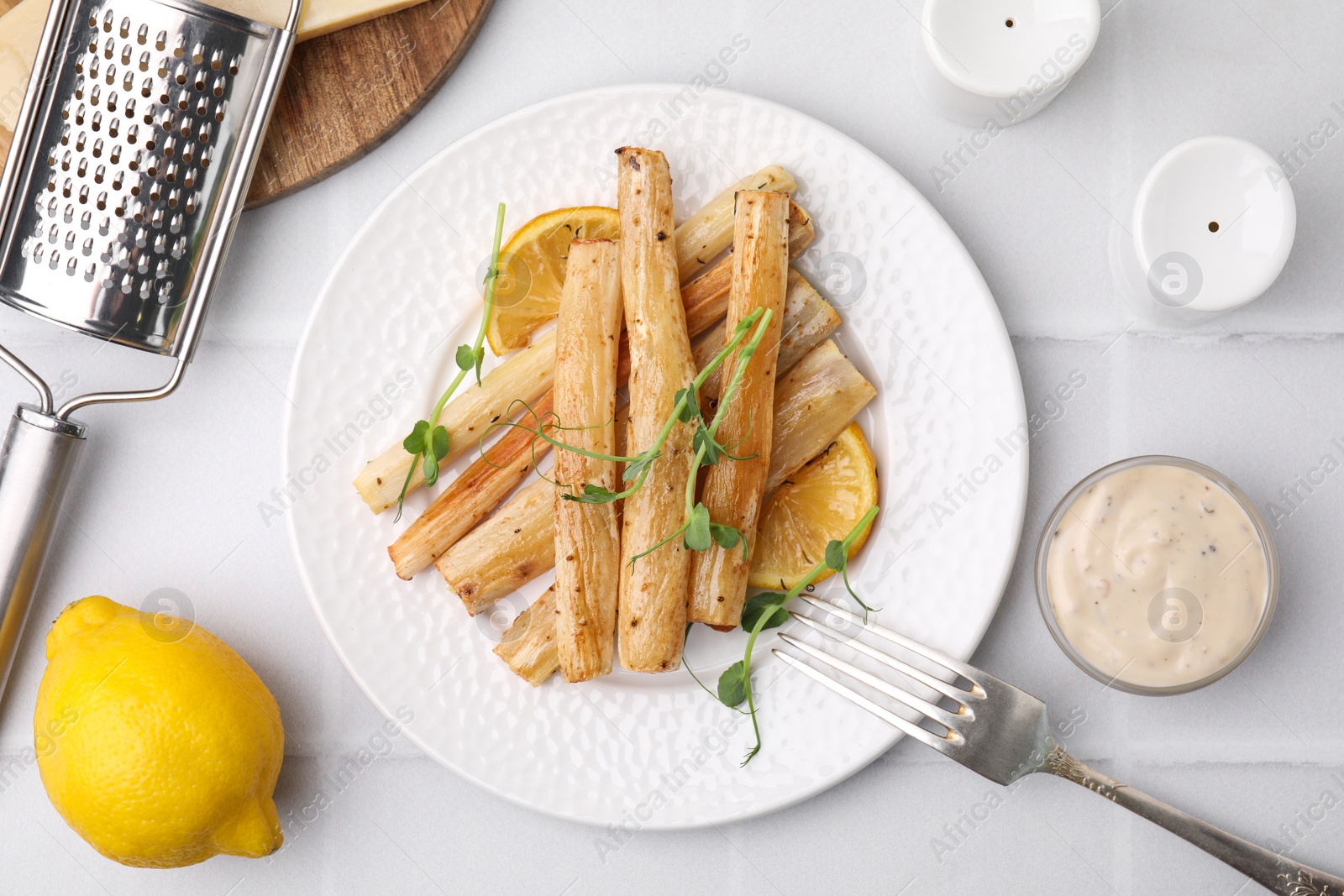 Photo of Plate with baked salsify roots, lemon, fork and sauce on white tiled table, flat lay