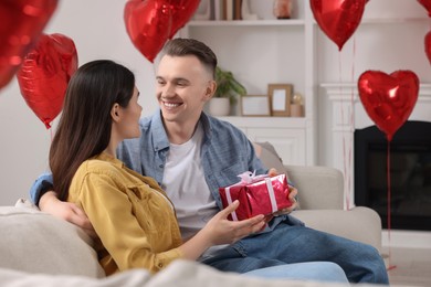 Happy couple celebrating Valentine's day. Beloved woman with gift box in room decorated with heart shaped air balloons