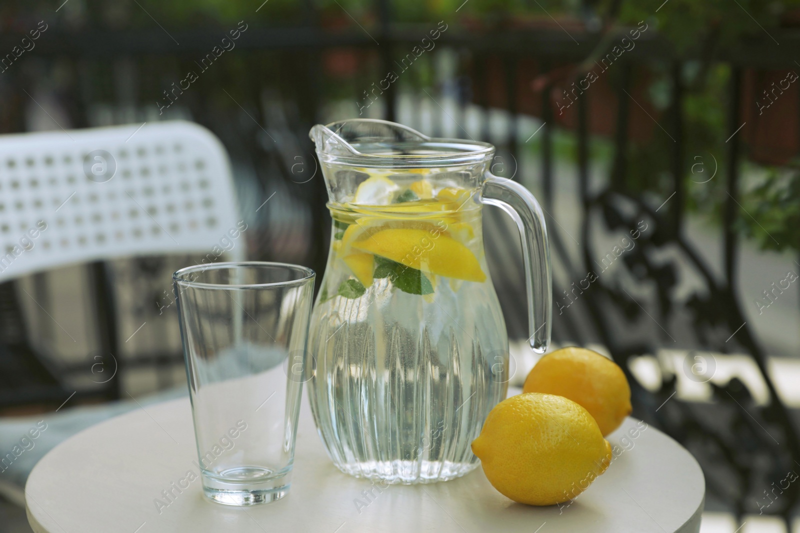 Photo of Jug with refreshing lemon water, glass and citrus fruits on light table outdoors
