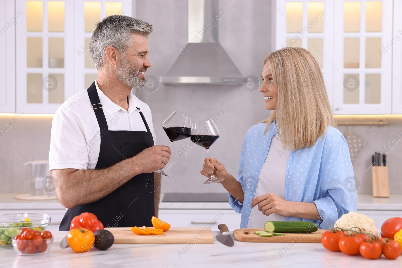 Photo of Happy affectionate couple with glasses of wine cooking together at white table in kitchen