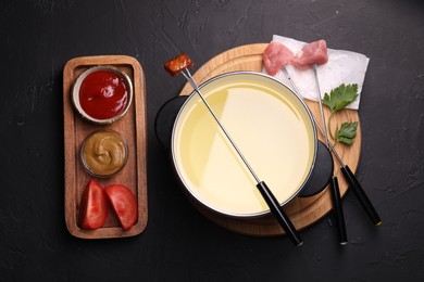 Photo of Fondue pot, forks with meat pieces and other products on black textured table, flat lay