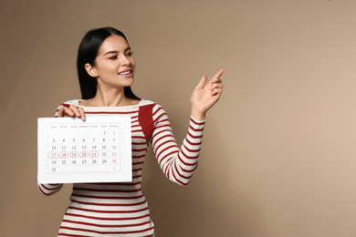 Young woman holding calendar with marked menstrual cycle days on beige background