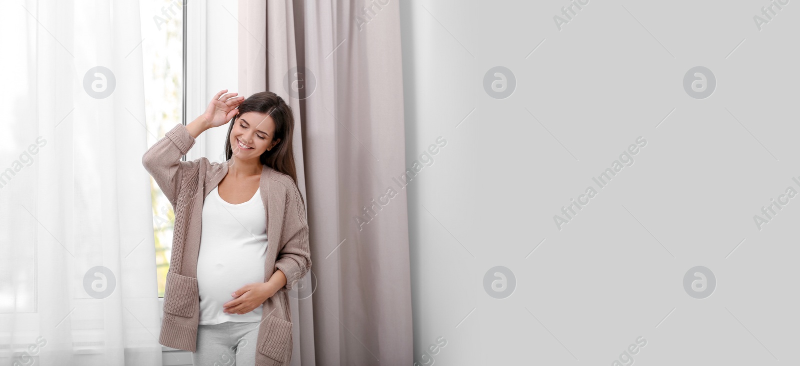 Image of Happy pregnant woman standing near window at home, space for text. Banner design
