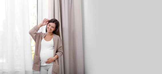 Image of Happy pregnant woman standing near window at home, space for text. Banner design
