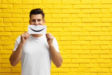 Photo of Man hiding emotions using card with drawn smile near yellow brick wall. Space for text