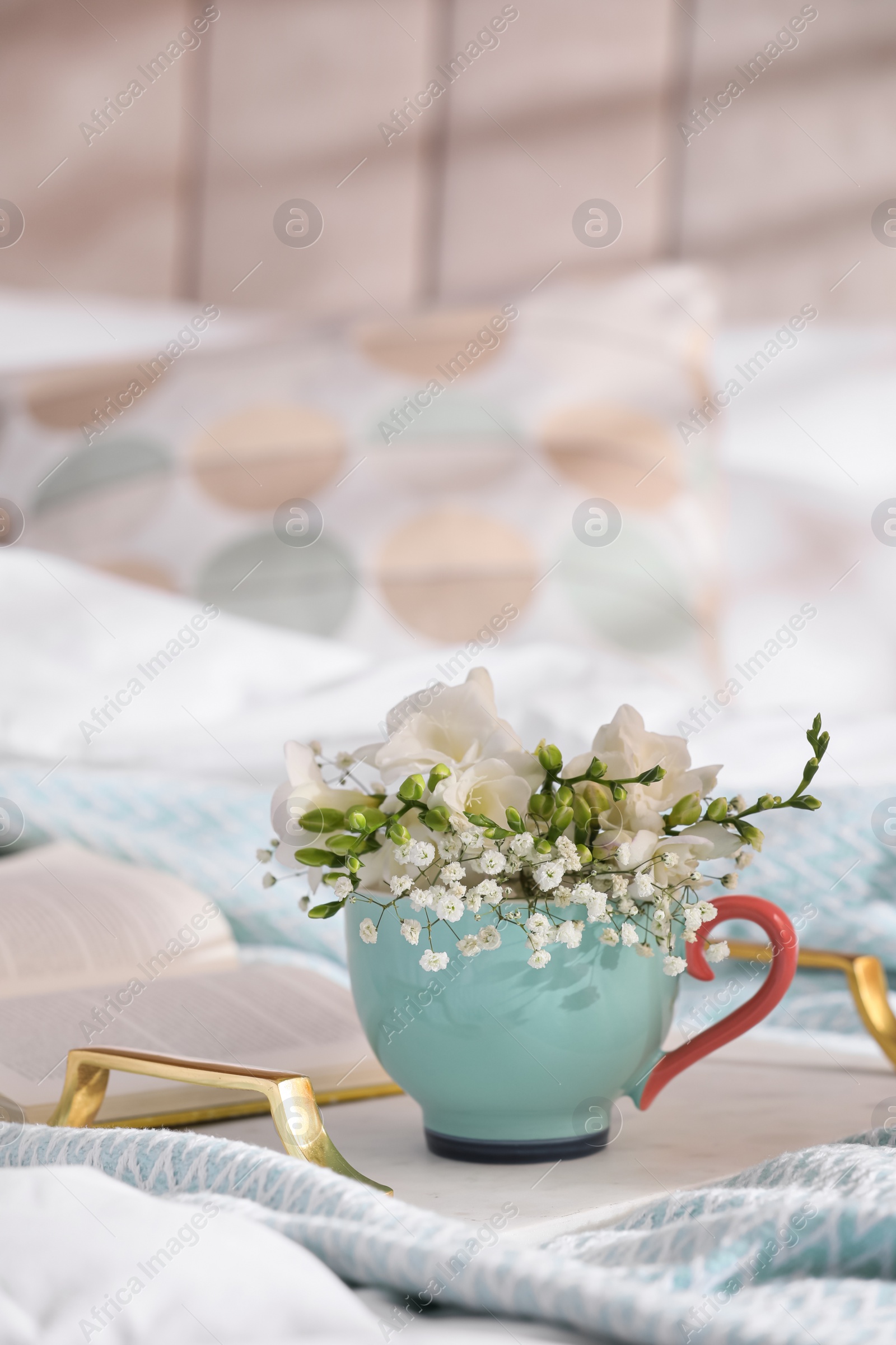 Photo of Beautiful bright flowers in cup, open book and tray on bed