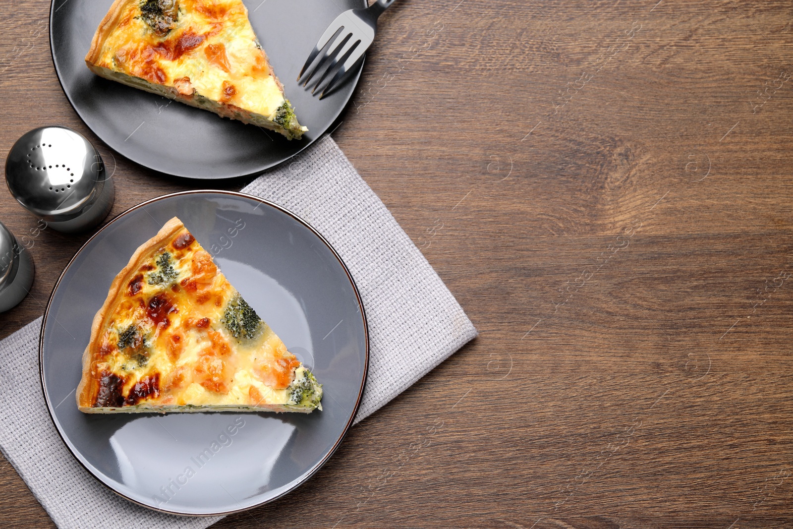 Photo of Pieces of delicious homemade salmon quiche with broccoli, fork and salt shaker on wooden table, flat lay. Space for text