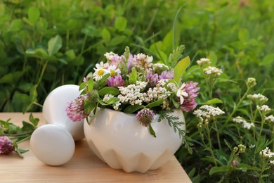 Ceramic mortar with pestle, different wildflowers and herbs on wooden board in meadow