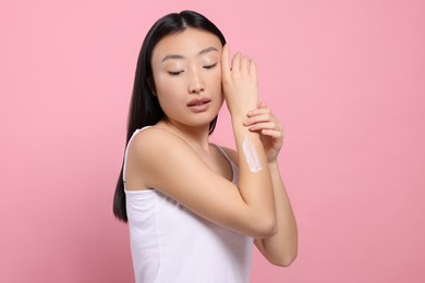 Photo of Beautiful young Asian woman applying body cream onto arm on pink background