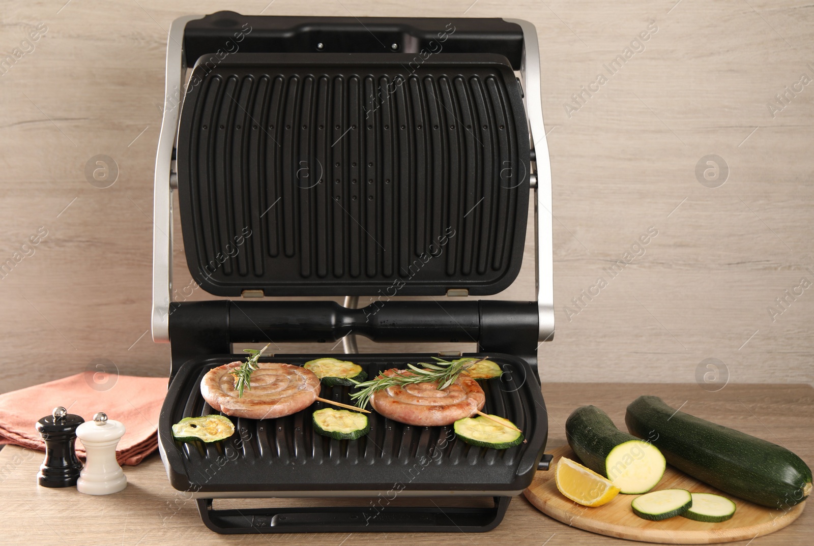 Photo of Electric grill with homemade sausages, zucchini and rosemary on wooden table